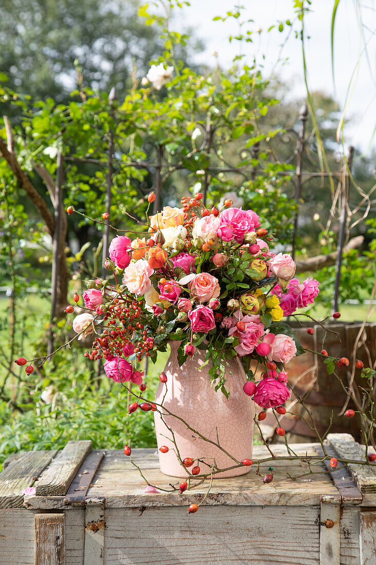 Rosenstrauß mit Hagebutten in rosa Keramikvase auf Holzkiste im Garten