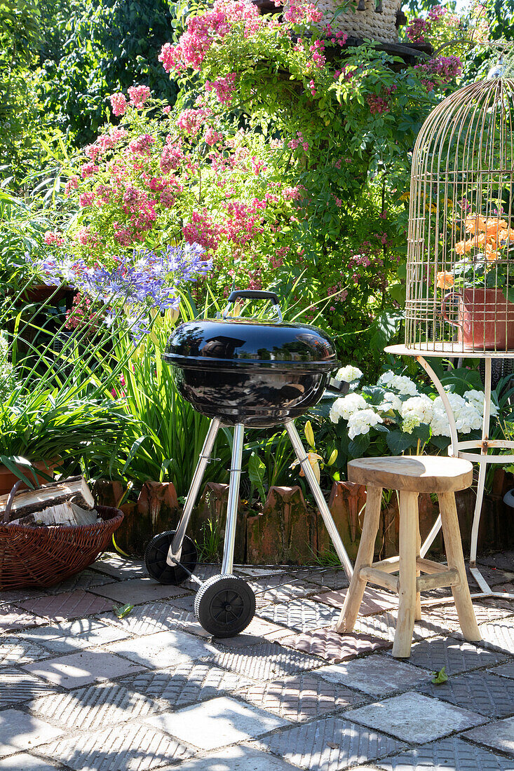 Kugelgrill im blühenden Garten neben rustikalem Hocker und Vogelkäfig