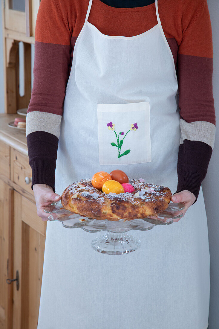 Person mit bestickter Schürze hält Kuchen mit bunten Ostereiern