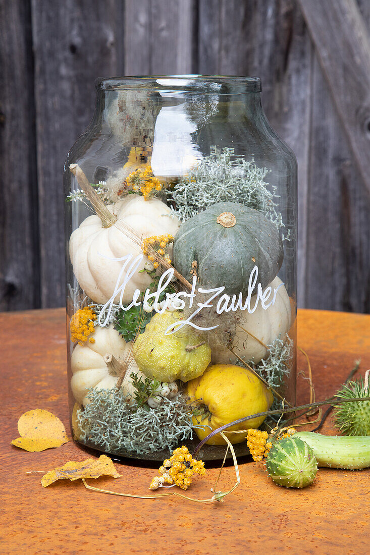 Decorative glass with Autumn magic lettering, filled with small pumpkins and dried flowers on a metal table