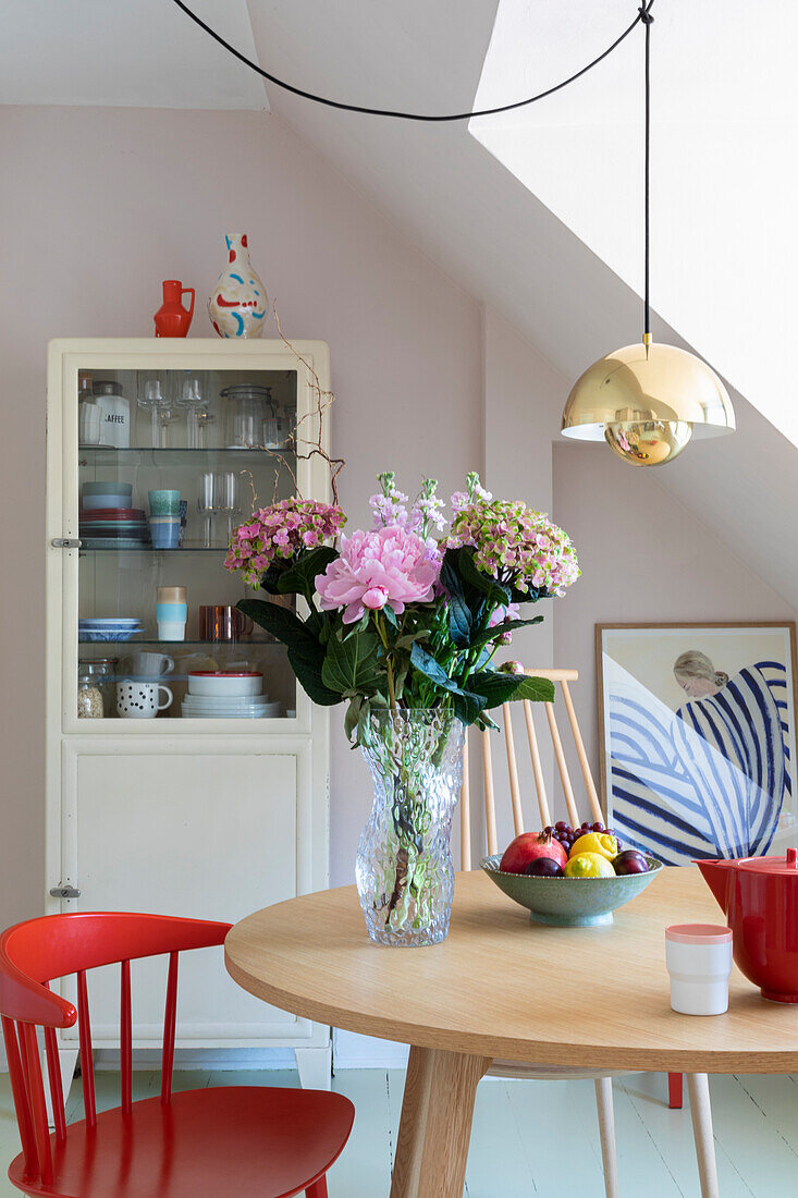 Round dining table with bouquet of flowers, red chair and wooden display cabinet in light-coloured room