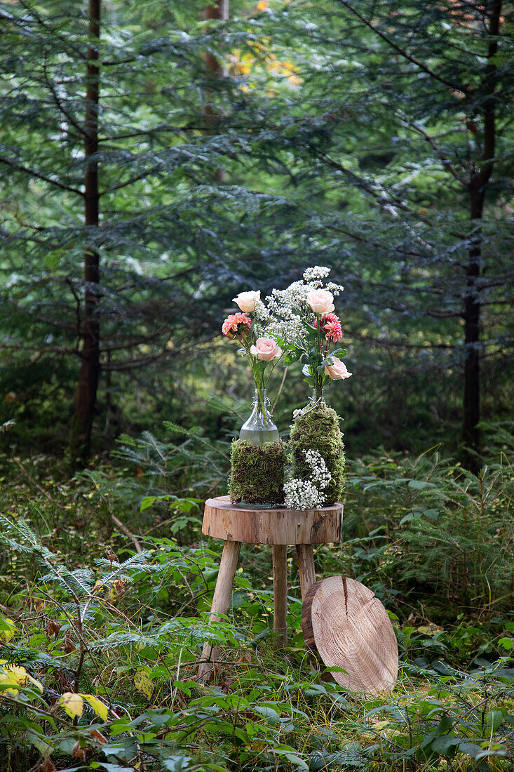 Glass bottles wrapped in moss with roses and gypsophila on a rustic wooden table in the forest