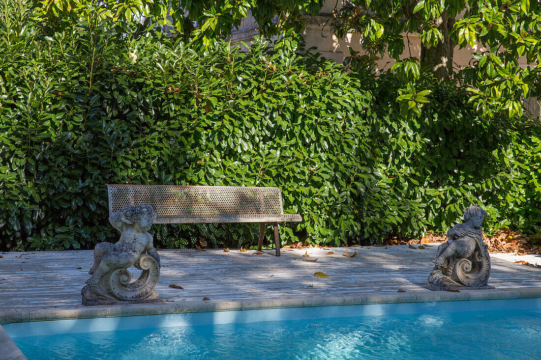 Steinbank und Wasserspeier-Statuen am Pool vor immergrüner Hecke