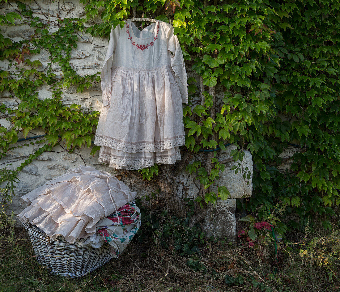 Weißes Vintage-Kleid an einer überwucherten Steinmauer, geflochtener Wäschekorb steht daneben