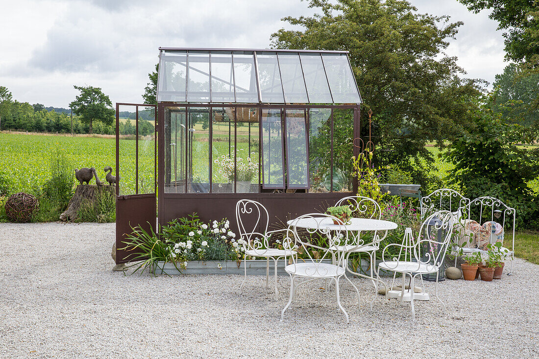 Romantic seating area with white iron furniture in front of greenhouse in the garden