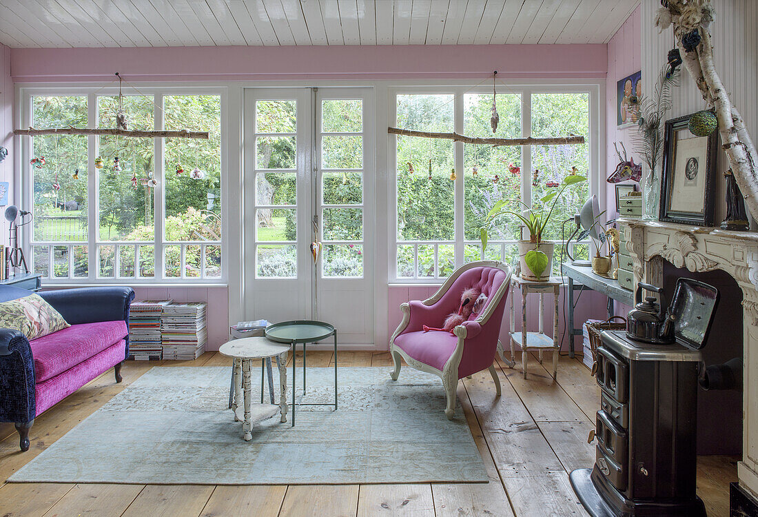 Living room with pink armchair and couch and wood-burning stove in front of large windows overlooking the garden