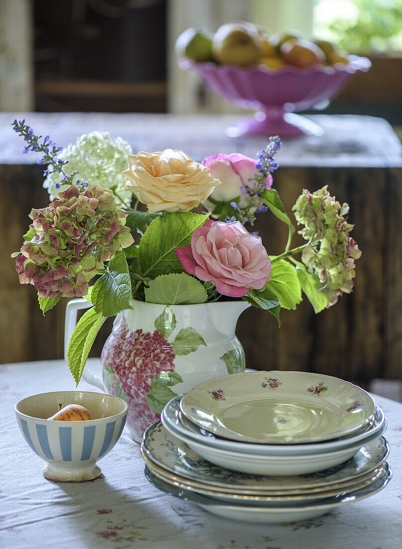 Blumenstrauß in Blumenvase auf gedecktem Tisch mit Geschirr und Obstschale im Hintergrund