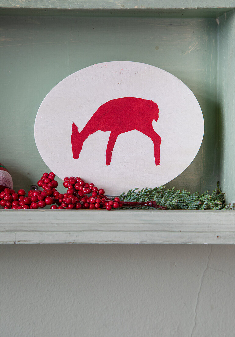 Hand-painted deer silhouette framed by red berries and evergreen branches on a wooden shelf