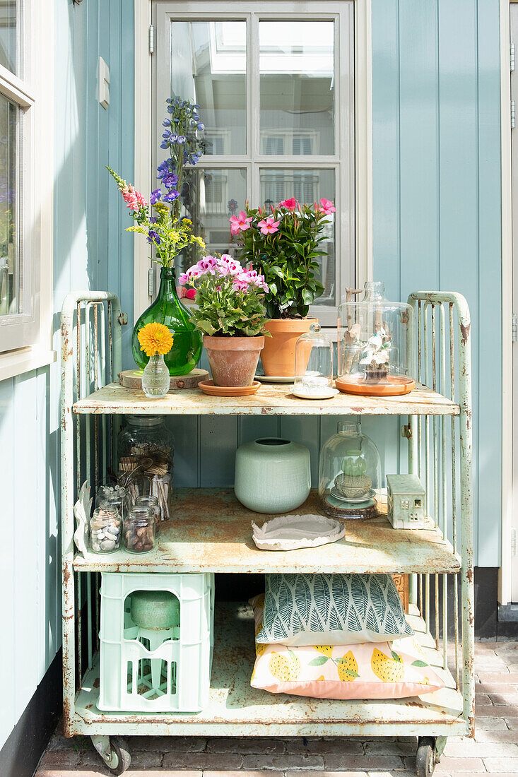 Blumenarrangement in Töpfen und Vasen auf einem Vintage-Rollwagen vor blau gestrichener Fassade