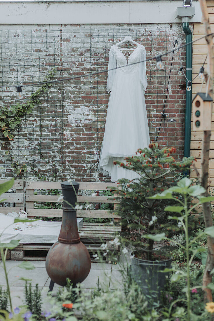 Brautkleid hängt an Gitter im Garten mit rostiger Vase und Pflanzen im Vordergrund