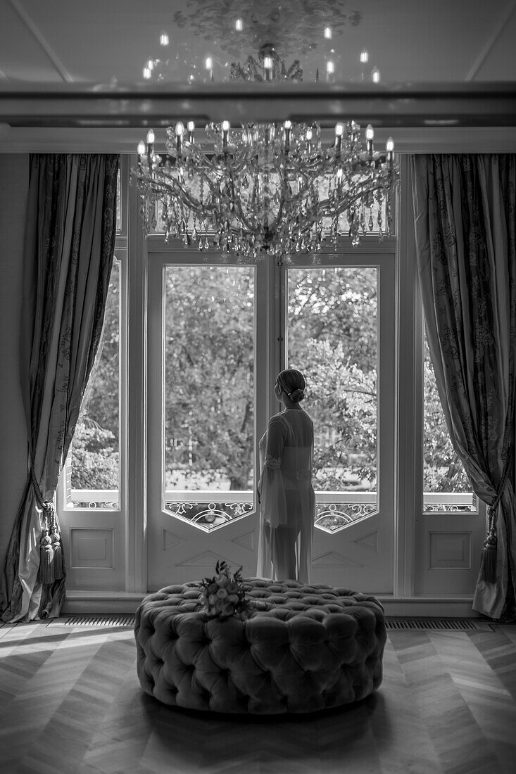 Woman in long dress looking out the window of a noble room with chandelier