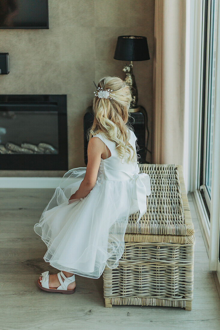 Girl in white festive dress sits on rattan bench in front of a fireplace