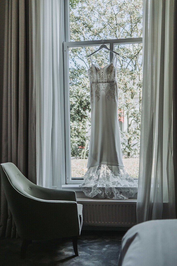 Brautkleid hängt am Fenster in Raum mit grünem Sessel und Gardinen