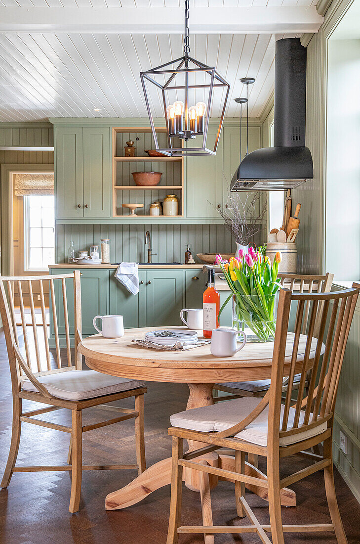 Round wooden table with bouquet of tulips and chairs in front of pastel green kitchen unit