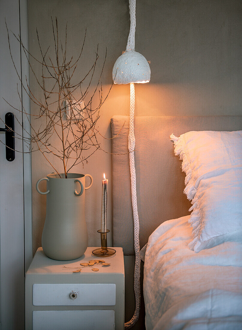 Bedside table with twigs in vase and candle next to bed and hanging lamp