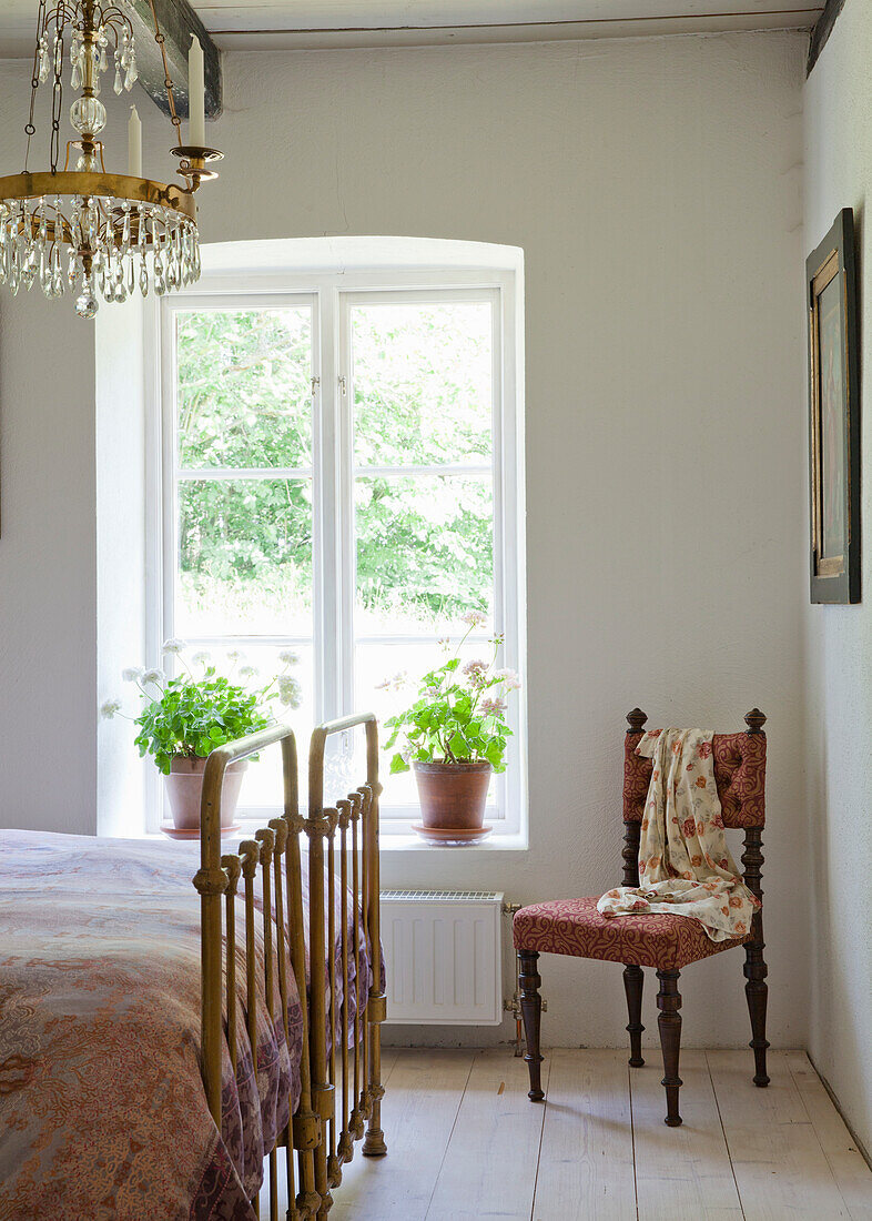 Bright bedroom with brass bed, antique chair, crystal chandelier and potted plants by the window