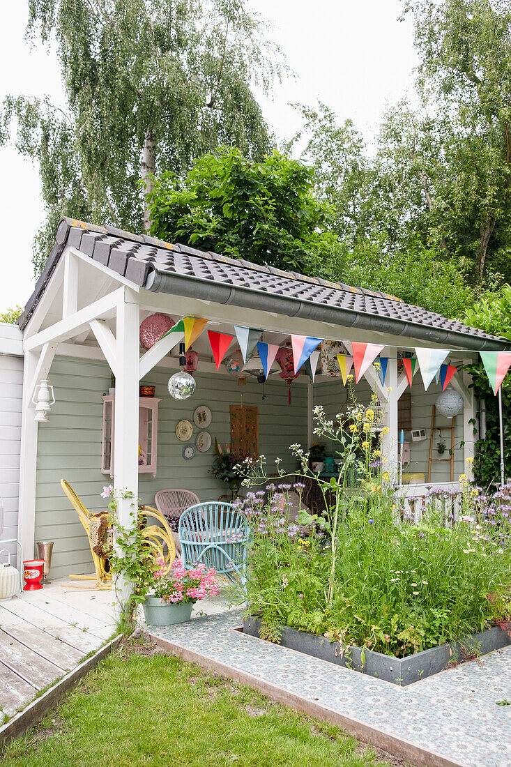 Farbenfrohes Gartenhaus mit Wimpelketten und Blumenbeet auf gefliester Terrasse