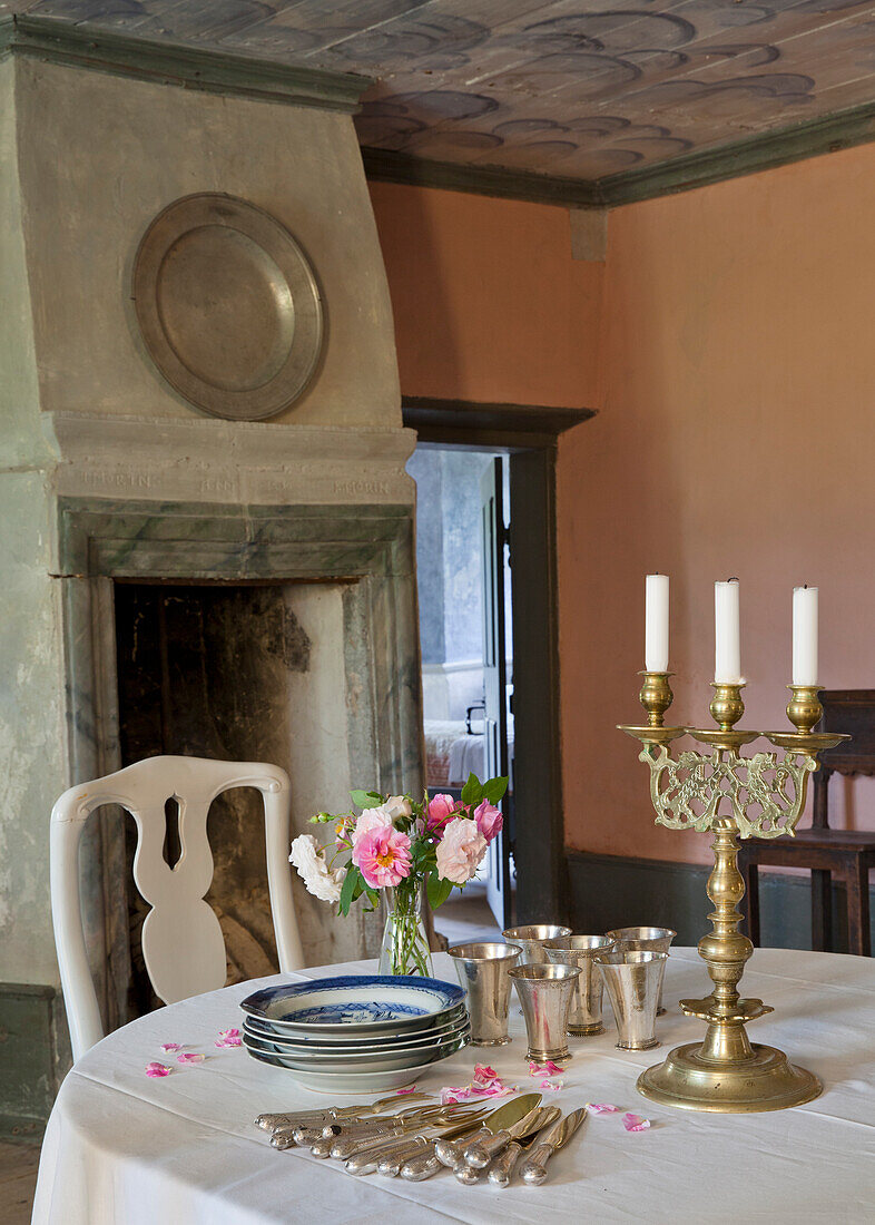 Round dining table with silver crockery, bouquet of flowers and candlesticks in front of antique fireplace