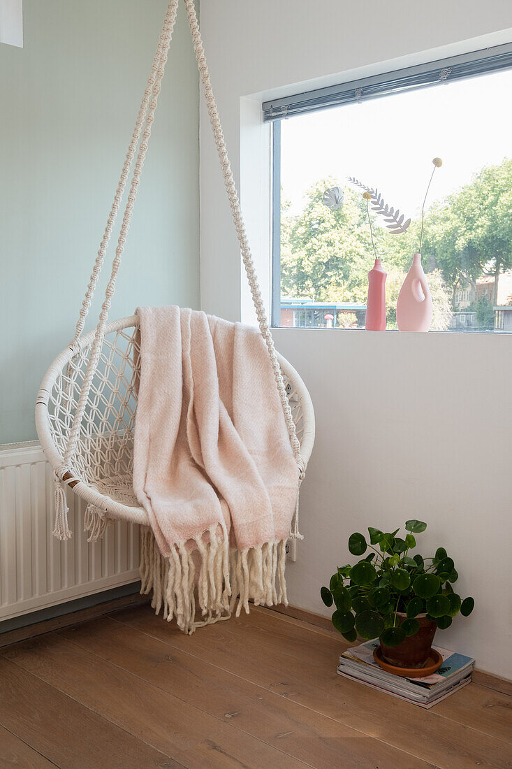 Hängesessel mit rosafarbener Decke vor Fenster und Zimmerpflanze