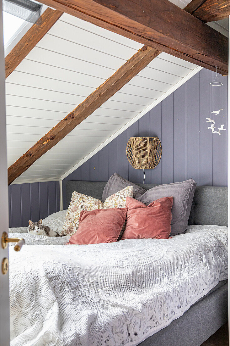 Bed in attic room with grey and pink cushions, wooden beams and wicker lamp