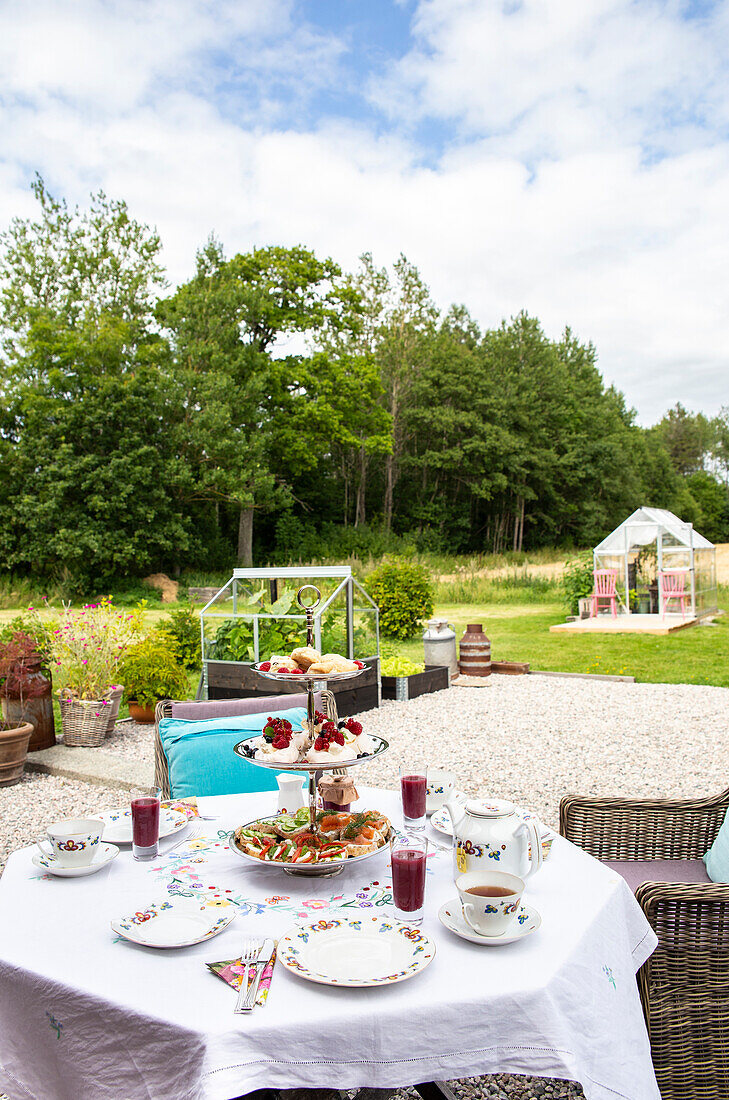 Laid garden table with porcelain crockery, etagere and teapot, background: greenhouse, trees and green meadow