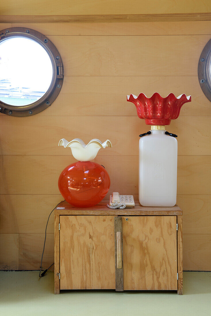 Two ornate table lamps on a wooden chest of drawers in front of porthole windows