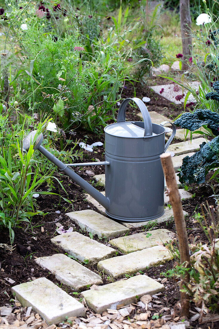 Gartenweg aus hellen Pflastersteinen mit grauer Gießkanne und Blumenbeete