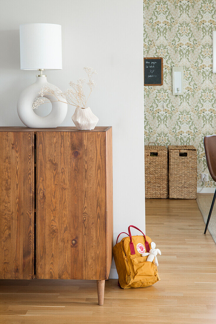 Holzkommode mit Trockenblumen und Lampe, gemusterte Tapete im Hintergrund