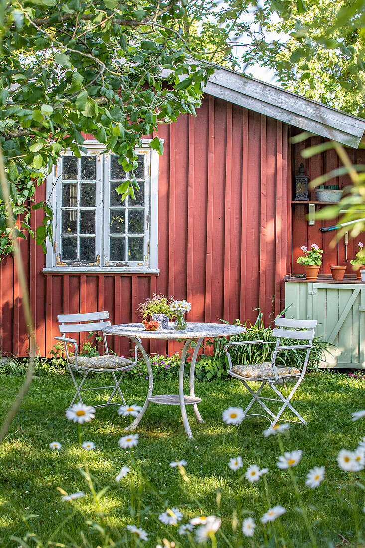 Gedeckter Gartentisch mit Blumenstrauß vor rotem Gartenhaus im Sommer