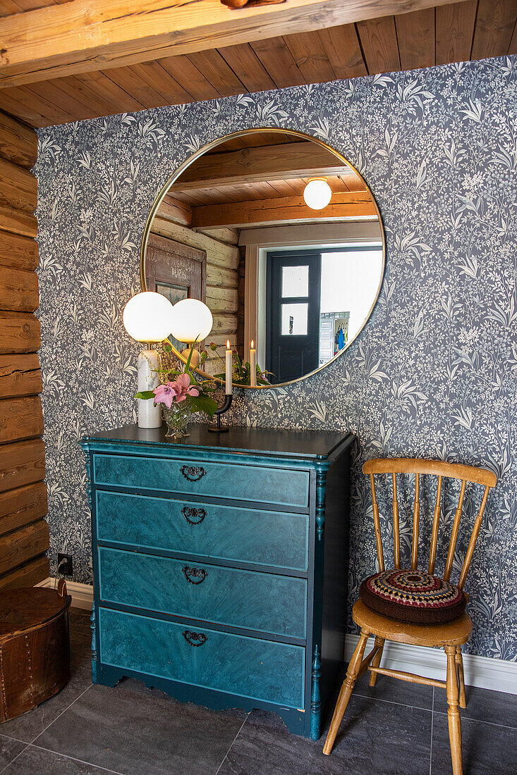Runder Wandspiegel über blauer Vintage-Kommode, daneben ein Holzstuhl