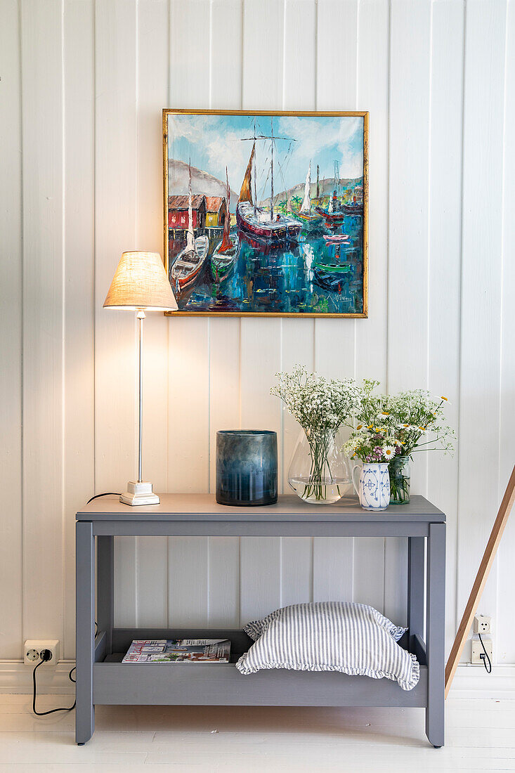 Scandinavian-style console table with fresh flowers and a lamp, painting with harbor scene above table
