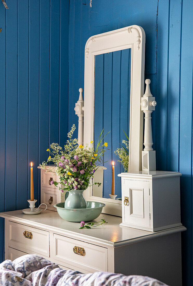 White dressing table with mirror and flower arrangement in front of blue wood paneling