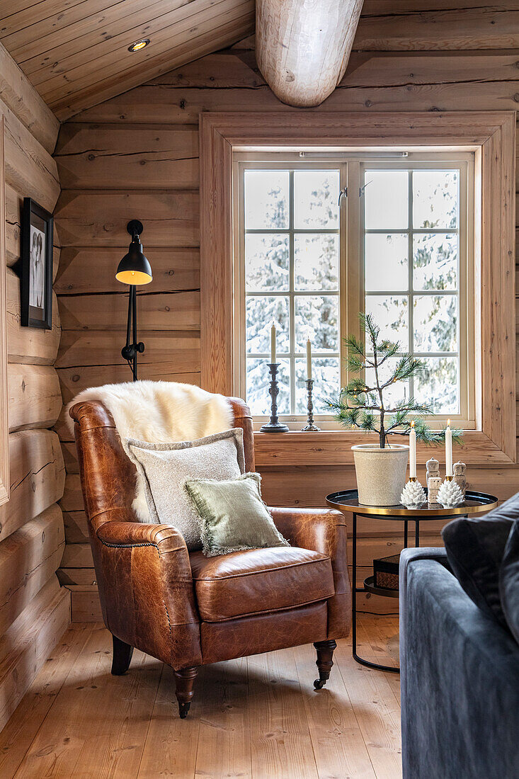 Comfortable leather armchair with fur and cushions in a rustic wooden house with a view of snow-covered trees