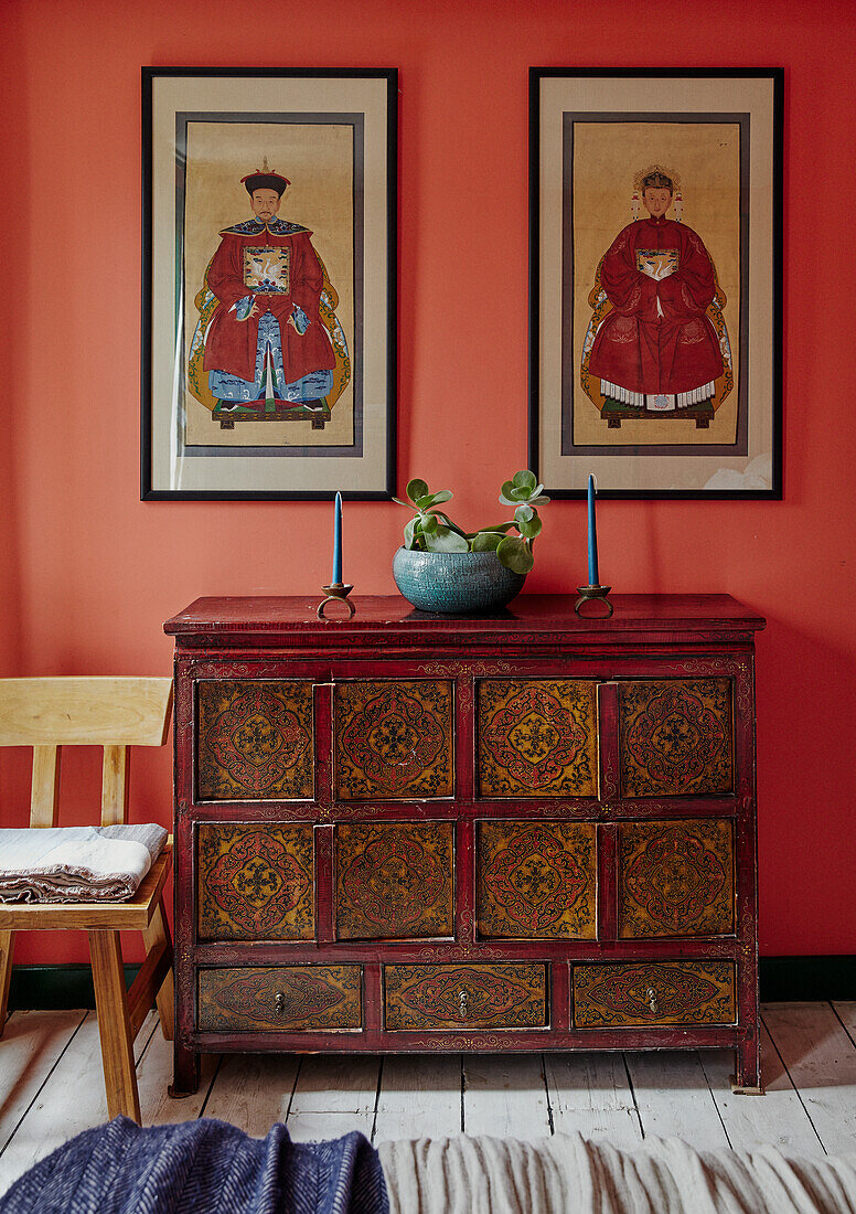 Antique painted wooden chest of drawers with Asian flair in front of orange wall with framed pictures