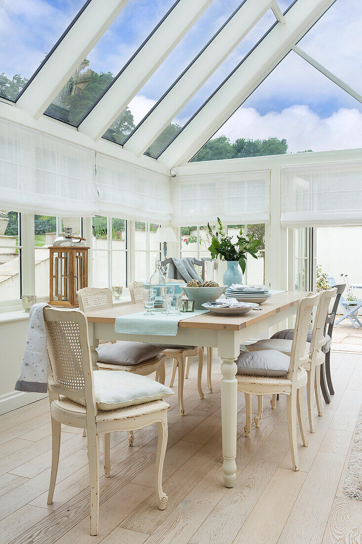 Dining area under a glass roof in the conservatory with light-colored wooden furniture