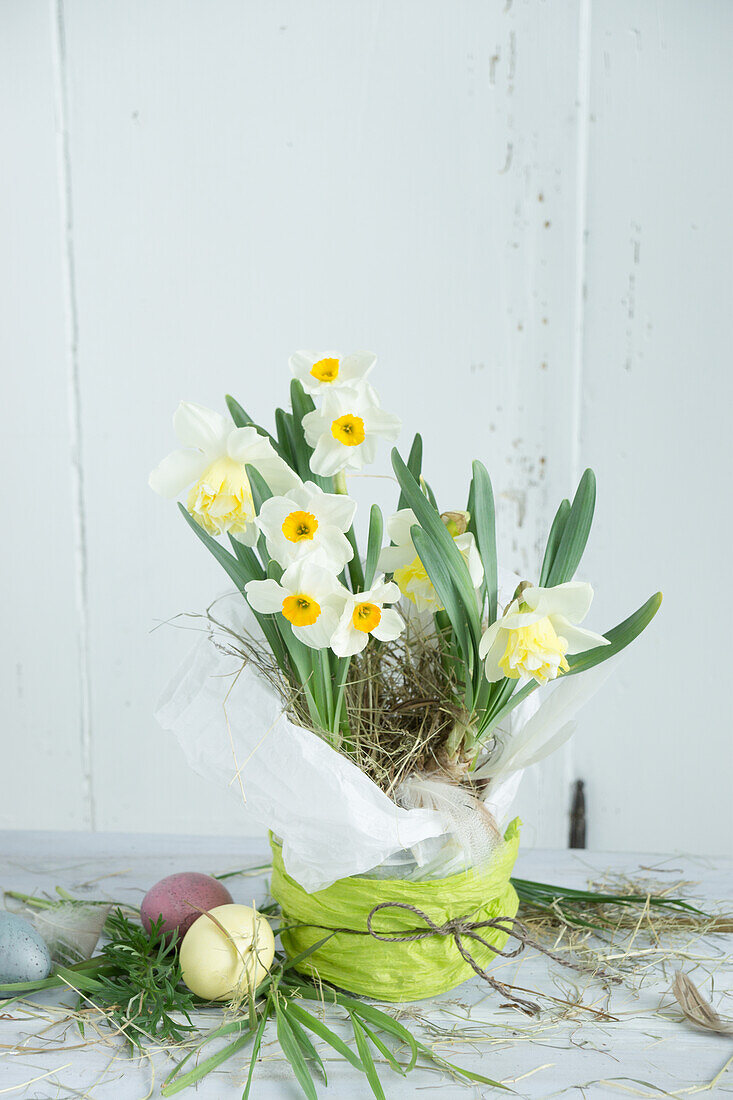 Daffodils in a flower pot with paper ribbon, next to it Easter eggs and straw decoration