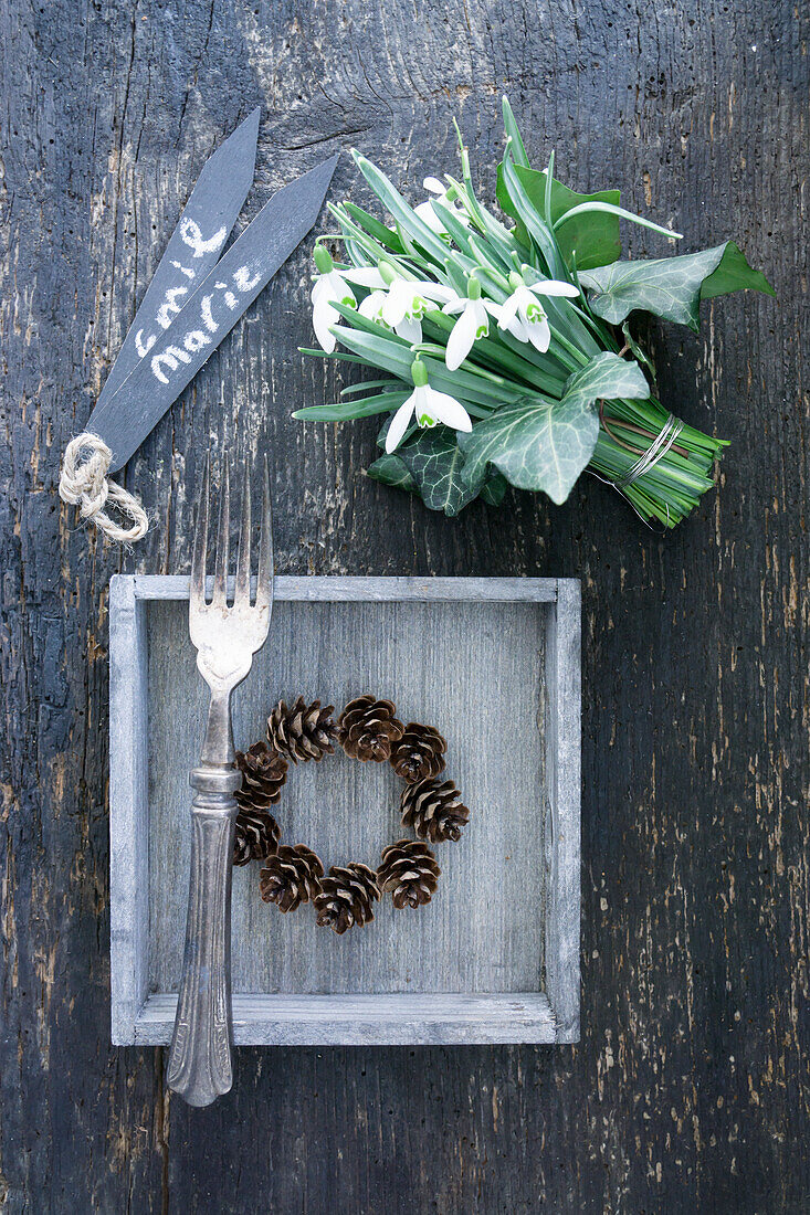 Wooden tray with wreath of larch cones and silver fork, bouquet of snowdrops with ivy leaves and name tag