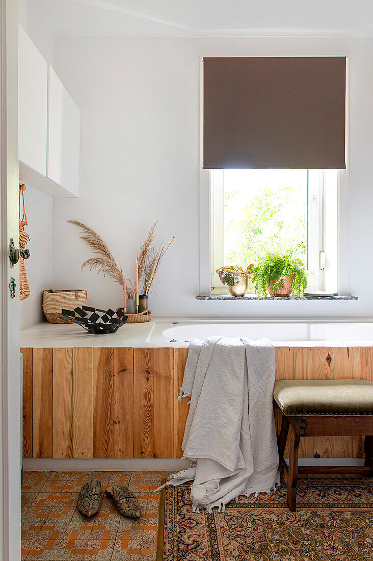 Bathroom with wooden bathtub paneling, plants by the window and stool with cushions