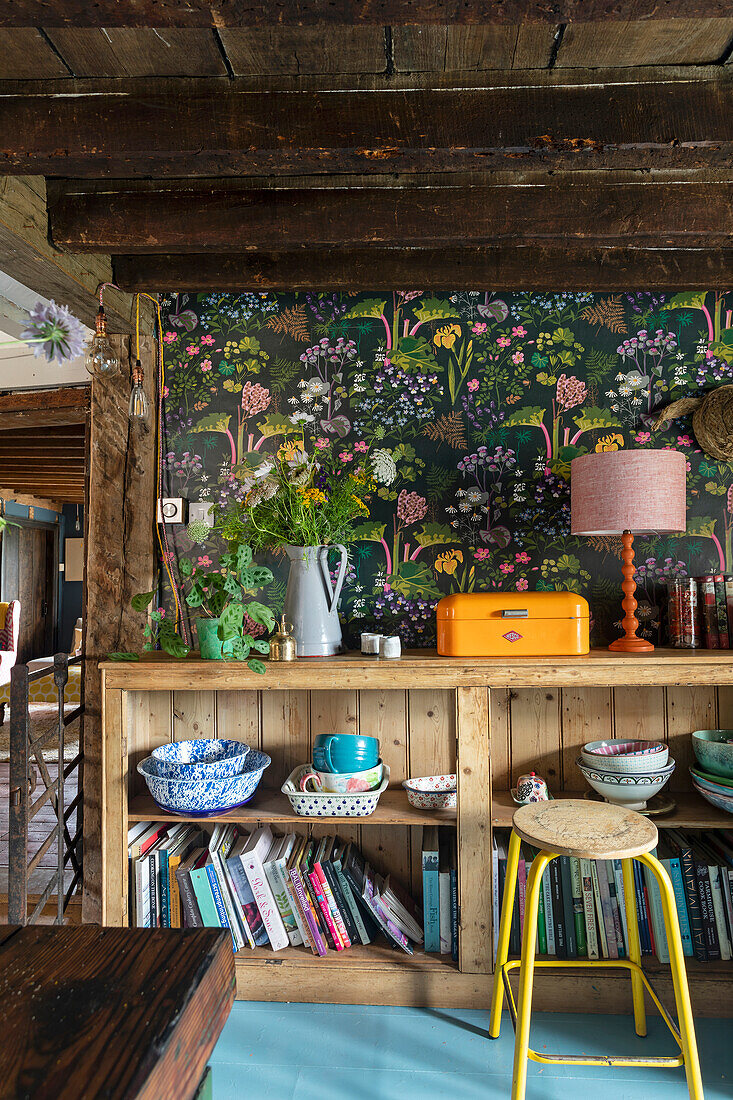 Shelf with colorful ceramics and books in front of floral wallpaper in rustic room