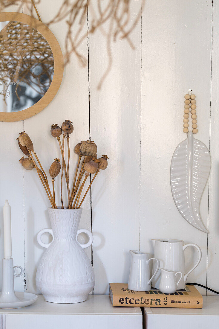 White vase with dried poppy capsules on white sideboard, decorative ceramic leaf as wall decoration