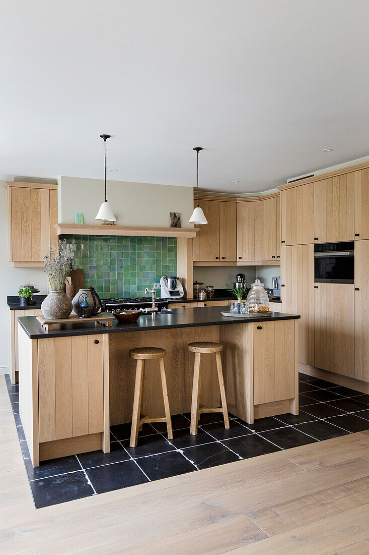 Kitchen with light-coloured wooden cupboards, green tiles, kitchen island and bar stools