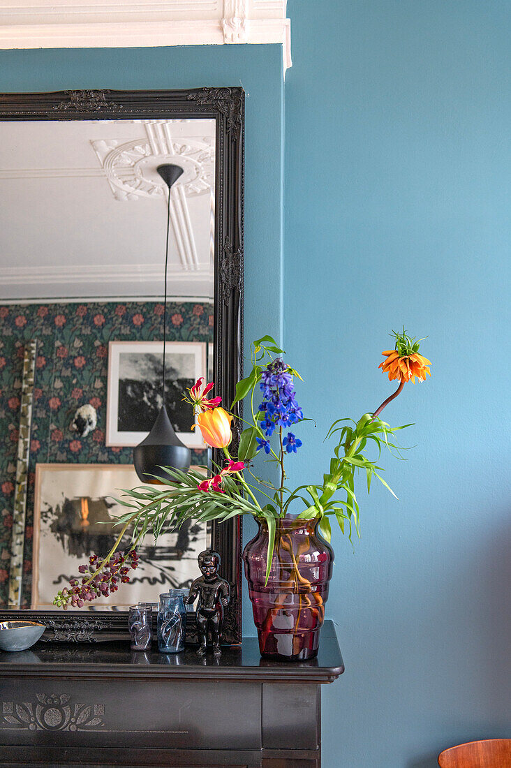 Buntes Blumenarrangement in roter Glasvase auf schwarzem Kaminsims vor blau gestrichener Wand