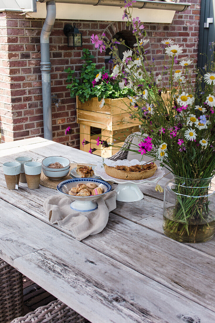 Holztisch im Freien mit Wildblumenstrauß, Kuchen und Gebäck