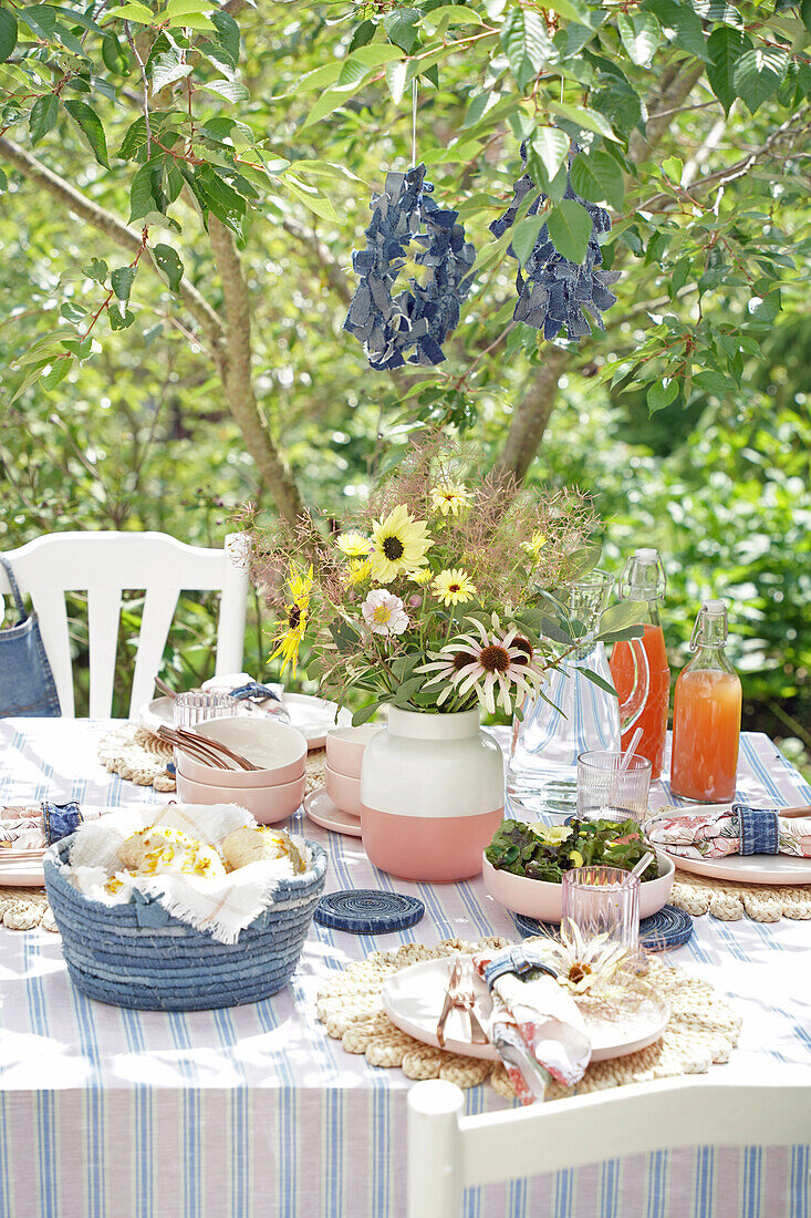 Laid garden table with bouquet of flowers and juice bottles under a tree