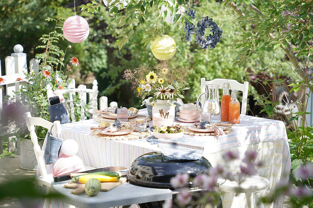 Gedeckter Gartentisch mit Sommerblumen und Saftflaschen unter Bäumen