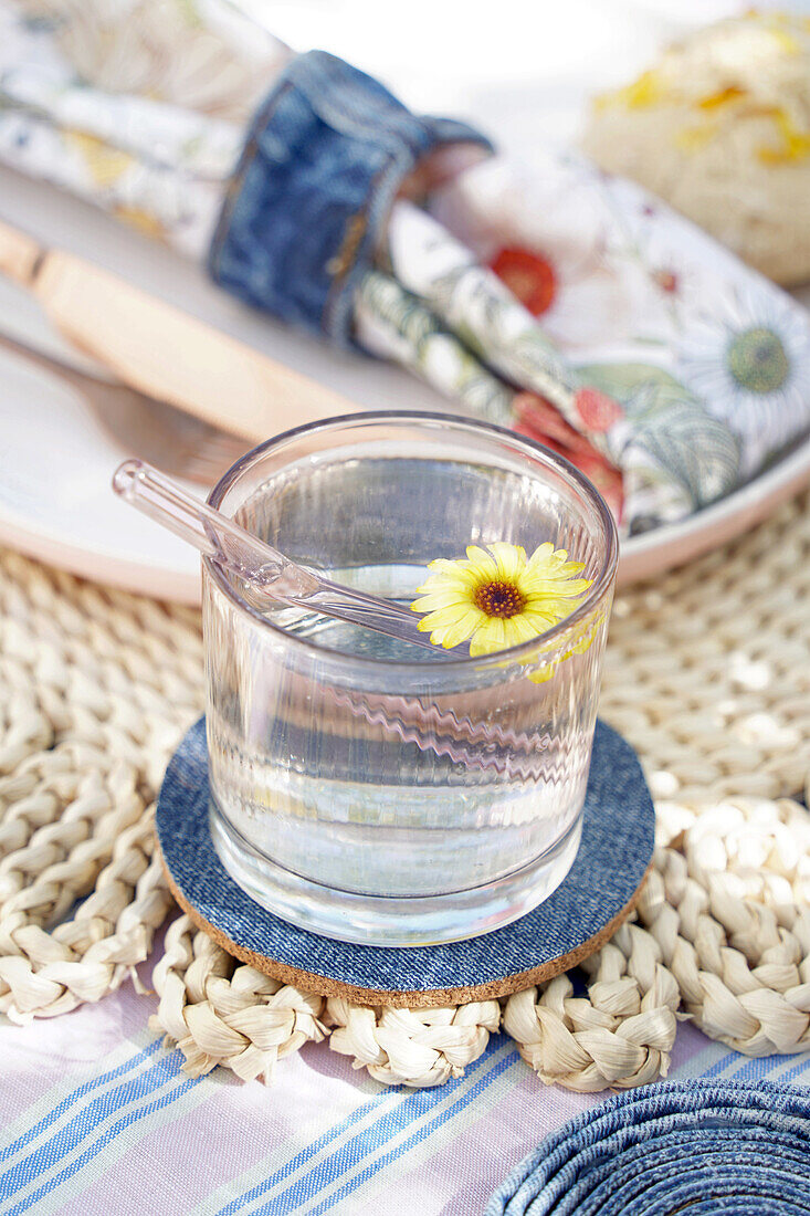 Water glass with yellow flower on wicker placemat and denim coaster