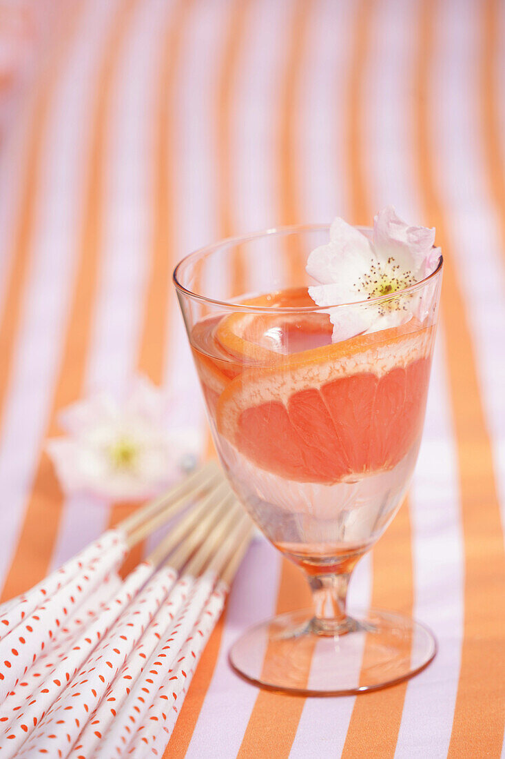 Glas mit Wasser, Blüte und Grapefruitscheiben auf gestreifter Tischdecke