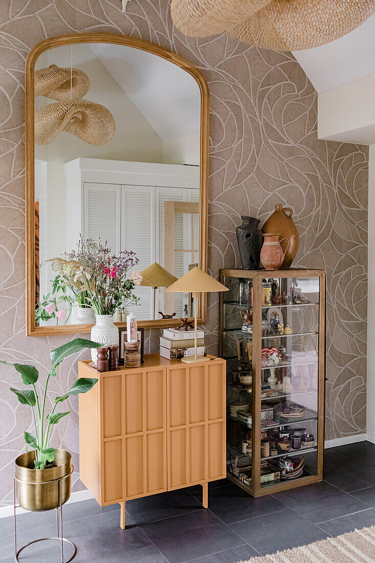 Sideboard and glass cabinet with decorative items in the hallway in front of patterned wallpaper and large mirror