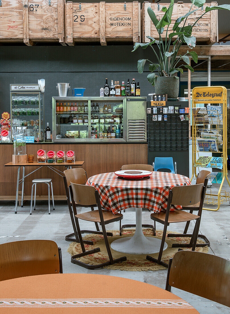 Table with checkered tablecloth and wooden chairs, sales counter, drinks and wooden crates