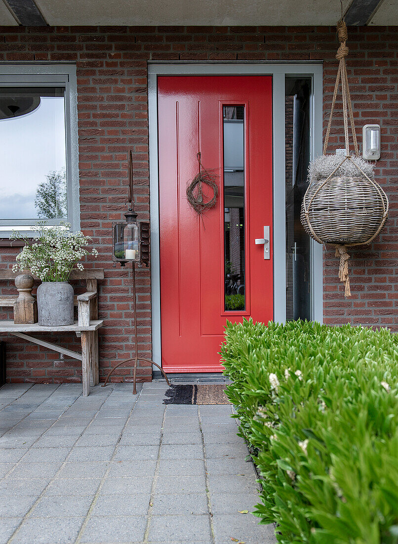 Red entrance door with narrow glass window and decorative wreath, wooden bench and large hanging basket